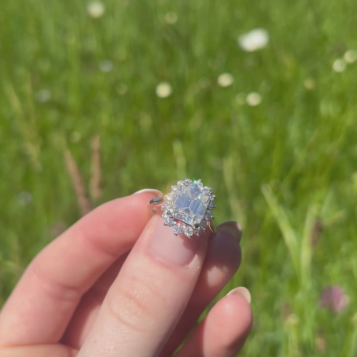 A beautiful continuum silver one and a three carat emerald shape moissanite halo ring, displaying the rainbow fire that is unique to this unique gemstone. This video has been shot in a field with wild flowers and you can hear the birds and wildlife in the back ground.